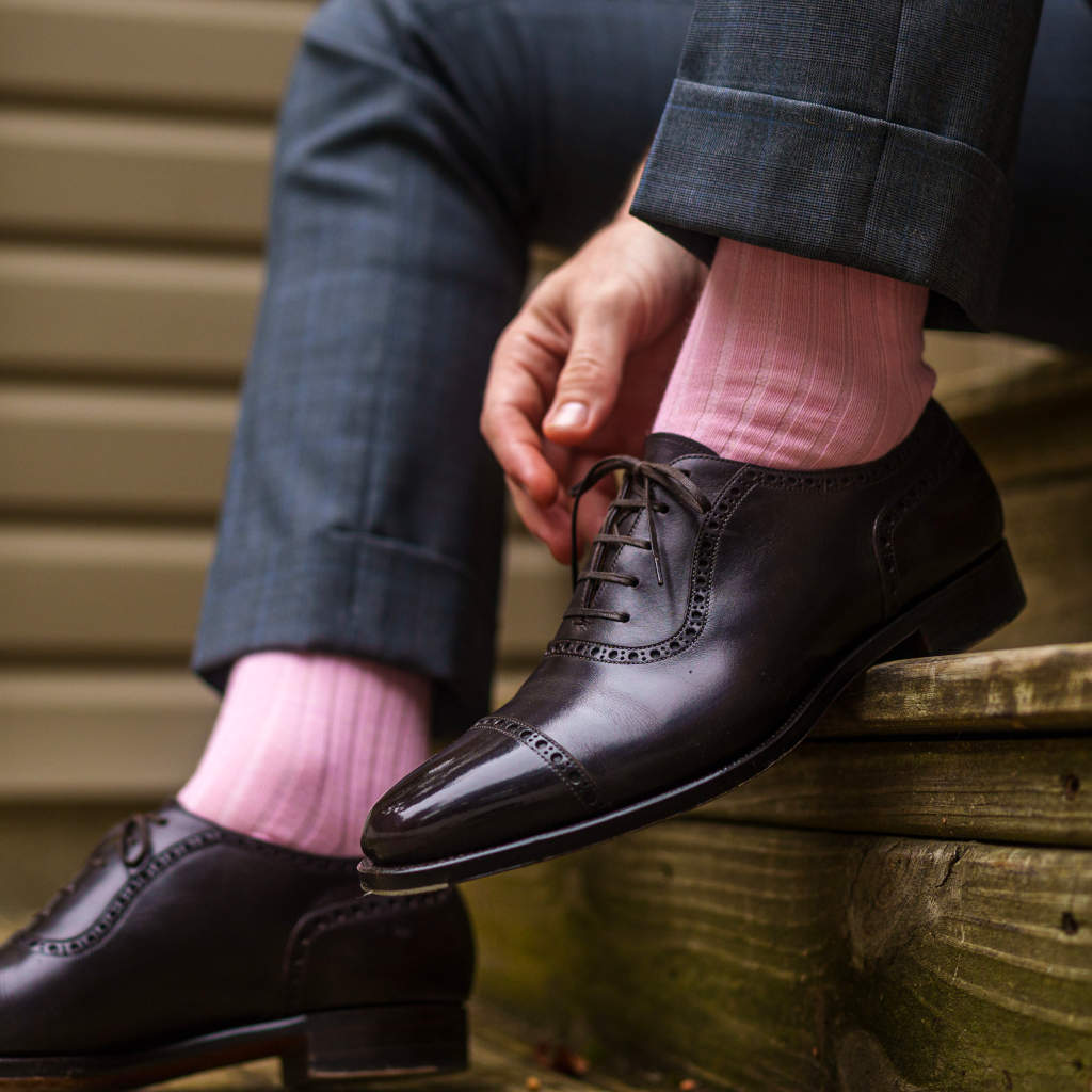 man wearing pink dress socks and grey trousers reaching down to adjust shoe laces on dark brown oxfords