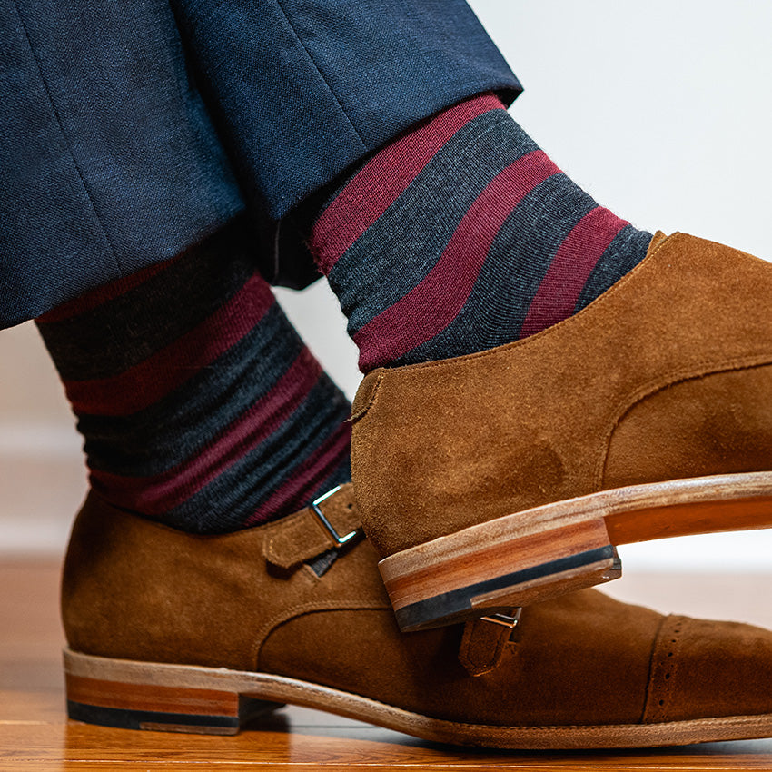man crossing ankles wearing burgundy and charcoal striped dress socks with a navy suit and light brown monkstraps