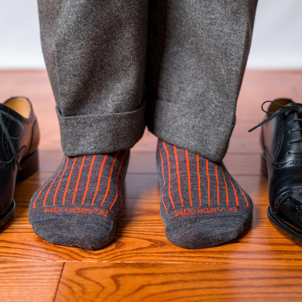 man standing on hardwood floor wearing grey slacks and orange and grey shadow stripe dress socks with BOARDROOM branding
