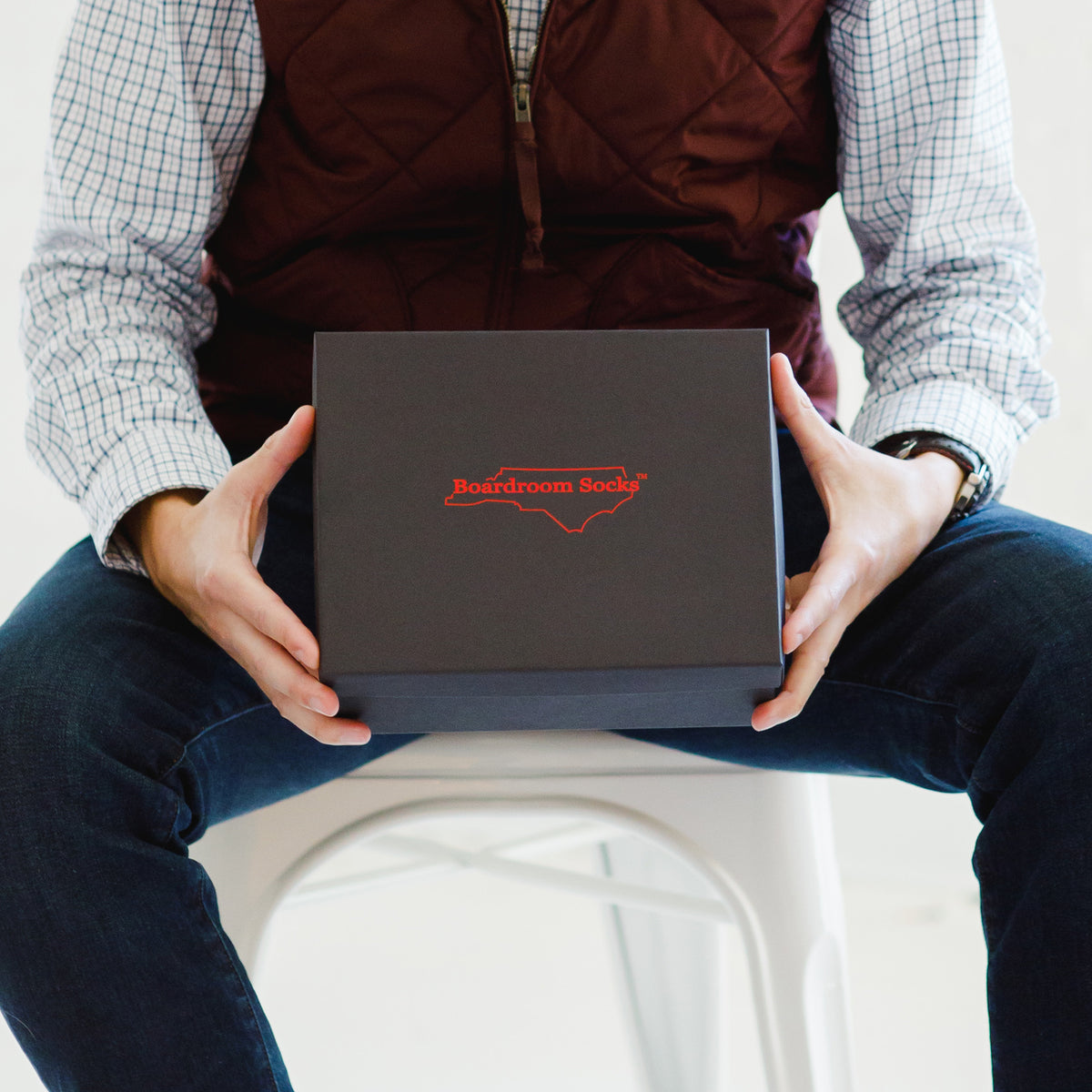 man seated on white chair holding black Boardroom Socks gift box filled with dress socks