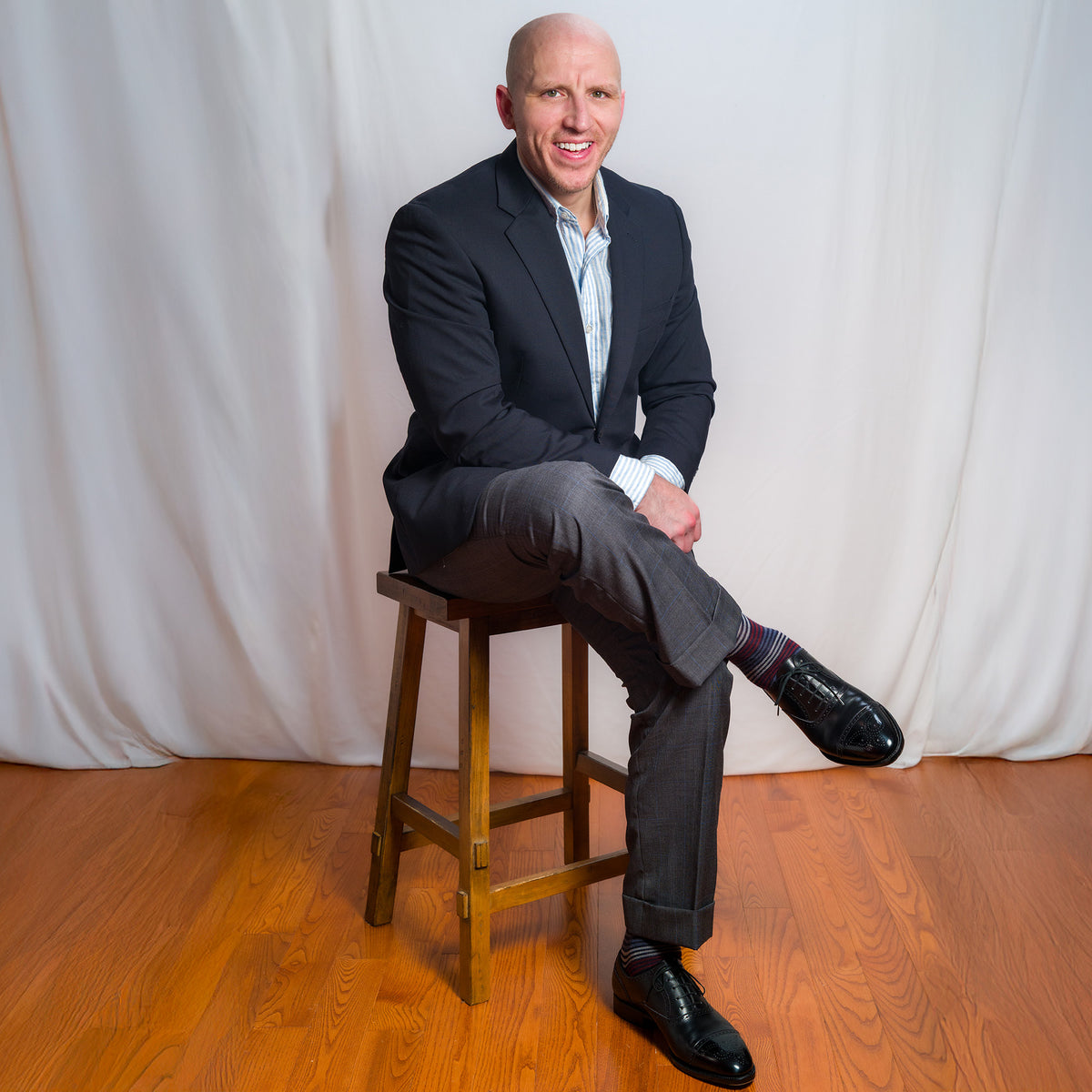 professional man in business attire wearing Boardroom Sockss while sitting on a wooden stool against a white curtain