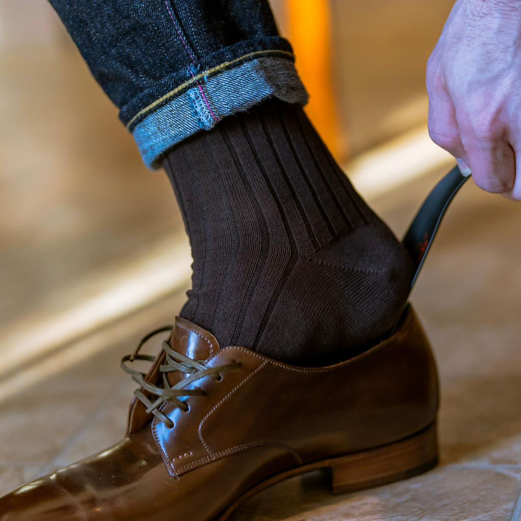 man wearing brown cotton dress socks using black metal shoe horn to put on dress shoes