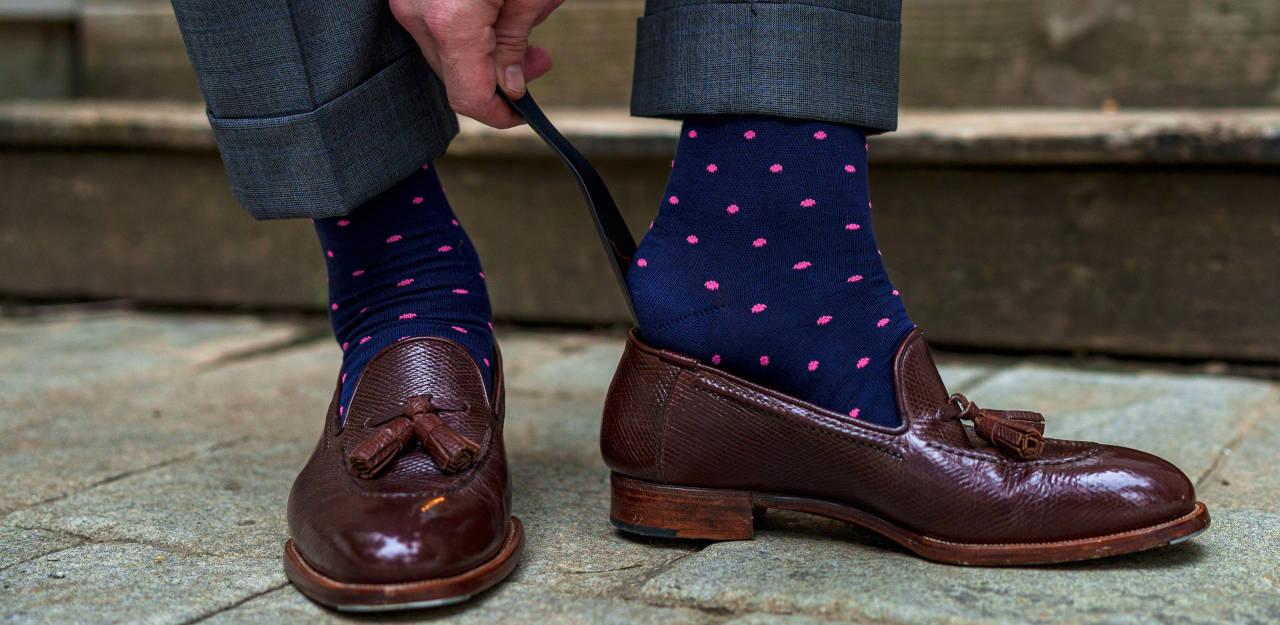 man wearing navy cotton dress socks using shoe horn to slide on tassel loafers