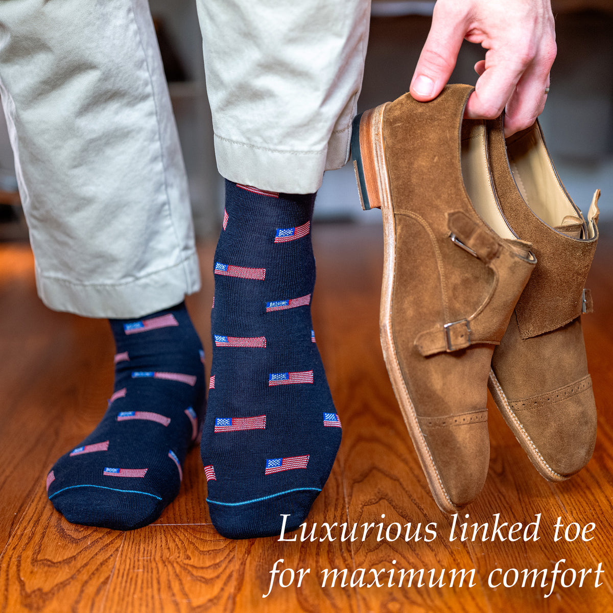 man walking on hardwood floor wearing American flag dress socks while holding brown suede monkstrap dress shoes