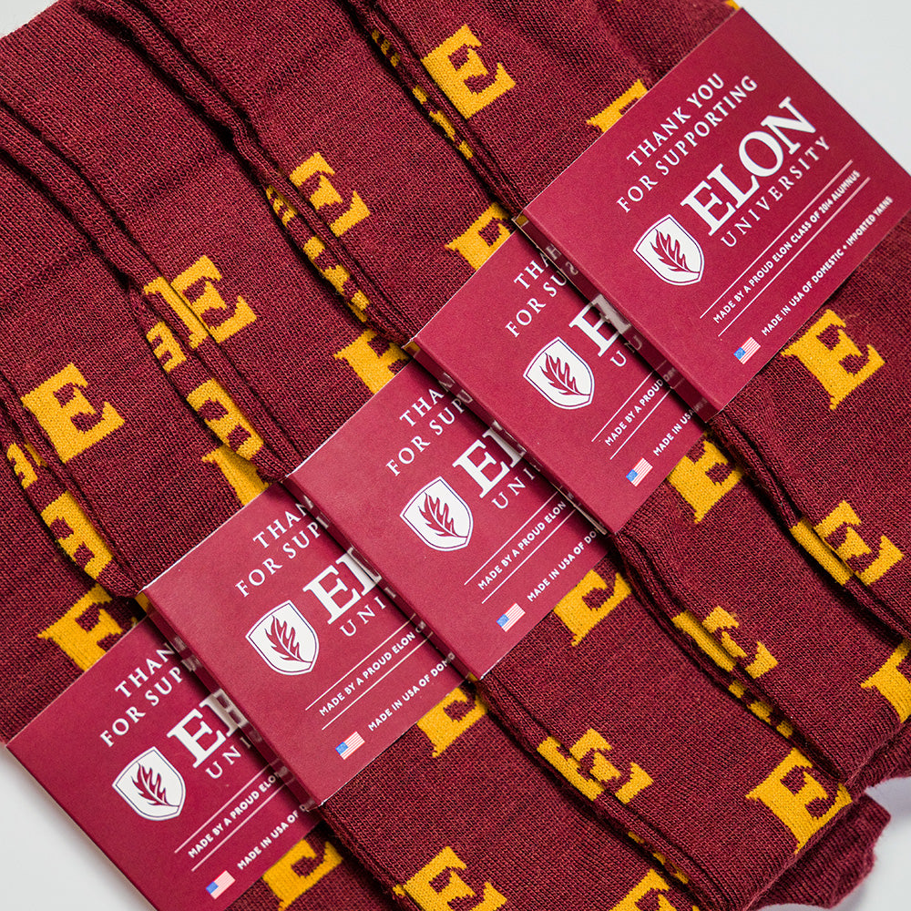 A close-up of several burgundy dress socks laid diagonally, each with a matching paper band. The socks have a repeating gold 'E' pattern, and the bands read 'THANK YOU FOR SUPPORTING ELON UNIVERSITY' with the university logo.