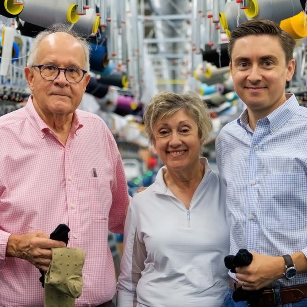 Three people standing together in a textile factory setting with spools of thread in the background, holding black knit socks. The man on the left wears a pink checkered shirt, the woman in the center wears a light blue polo, and the man on the right wears a light blue checkered shirt.