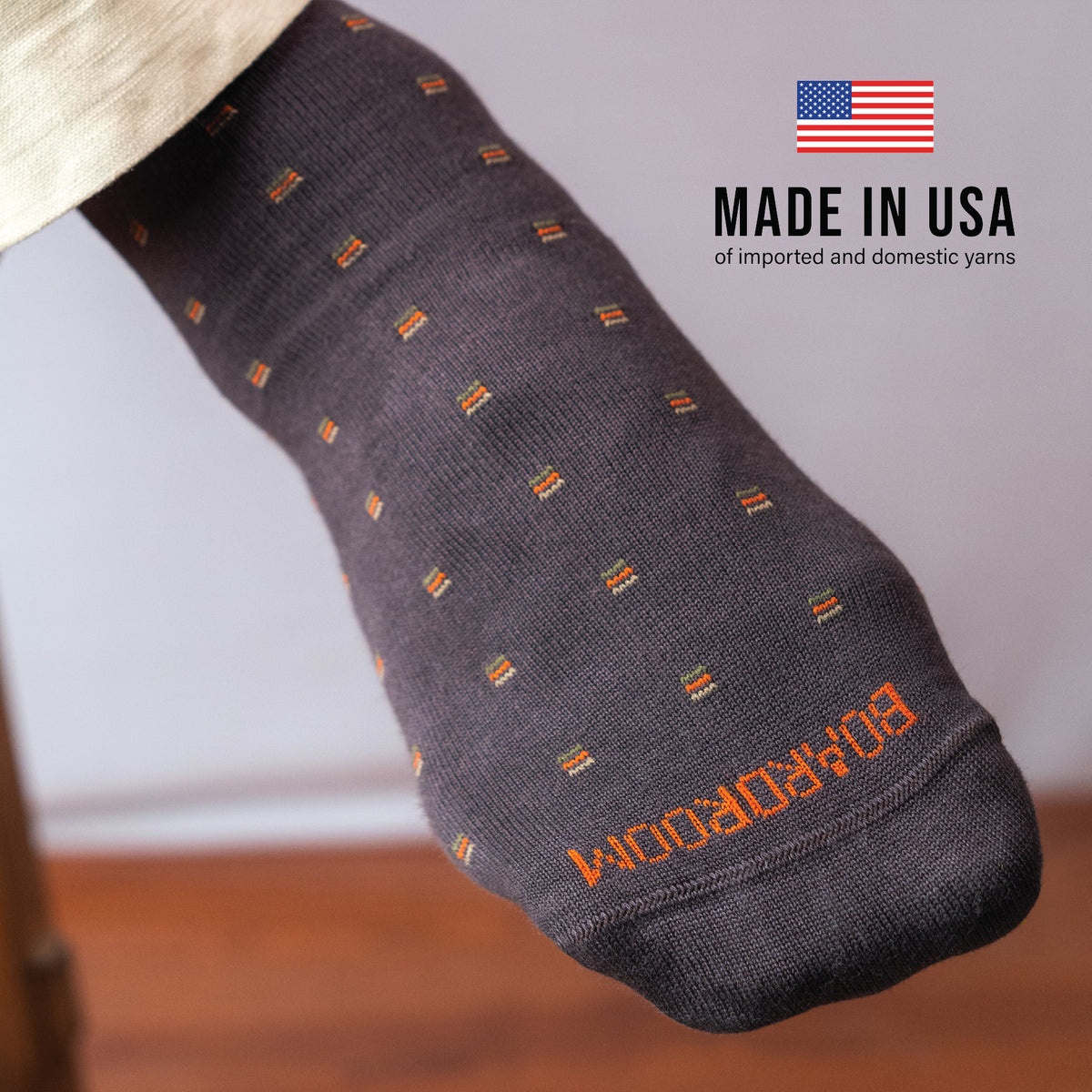 detailed image of brown patterned dress sock on foot worn with American flag image and text overlay about being made in the USA of imported and domestic yarns
