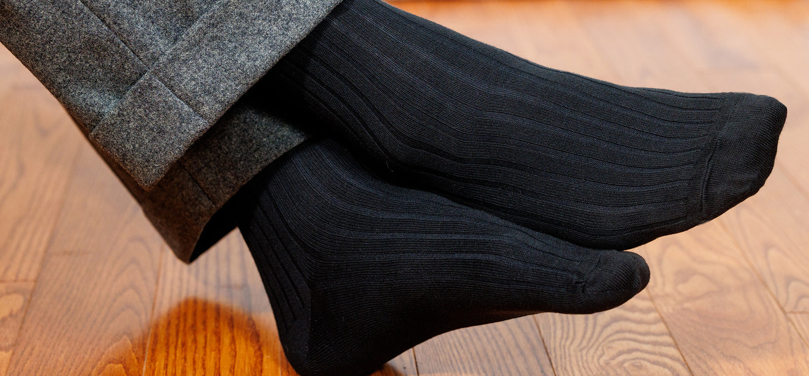 man wearing black ribbed dress socks with grey slacks crossing ankles on wood floor