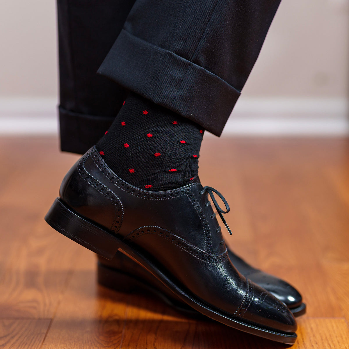 man crossing ankles wearing black wool dress socks decorated with red dots paired with charcoal slacks and black oxfords
