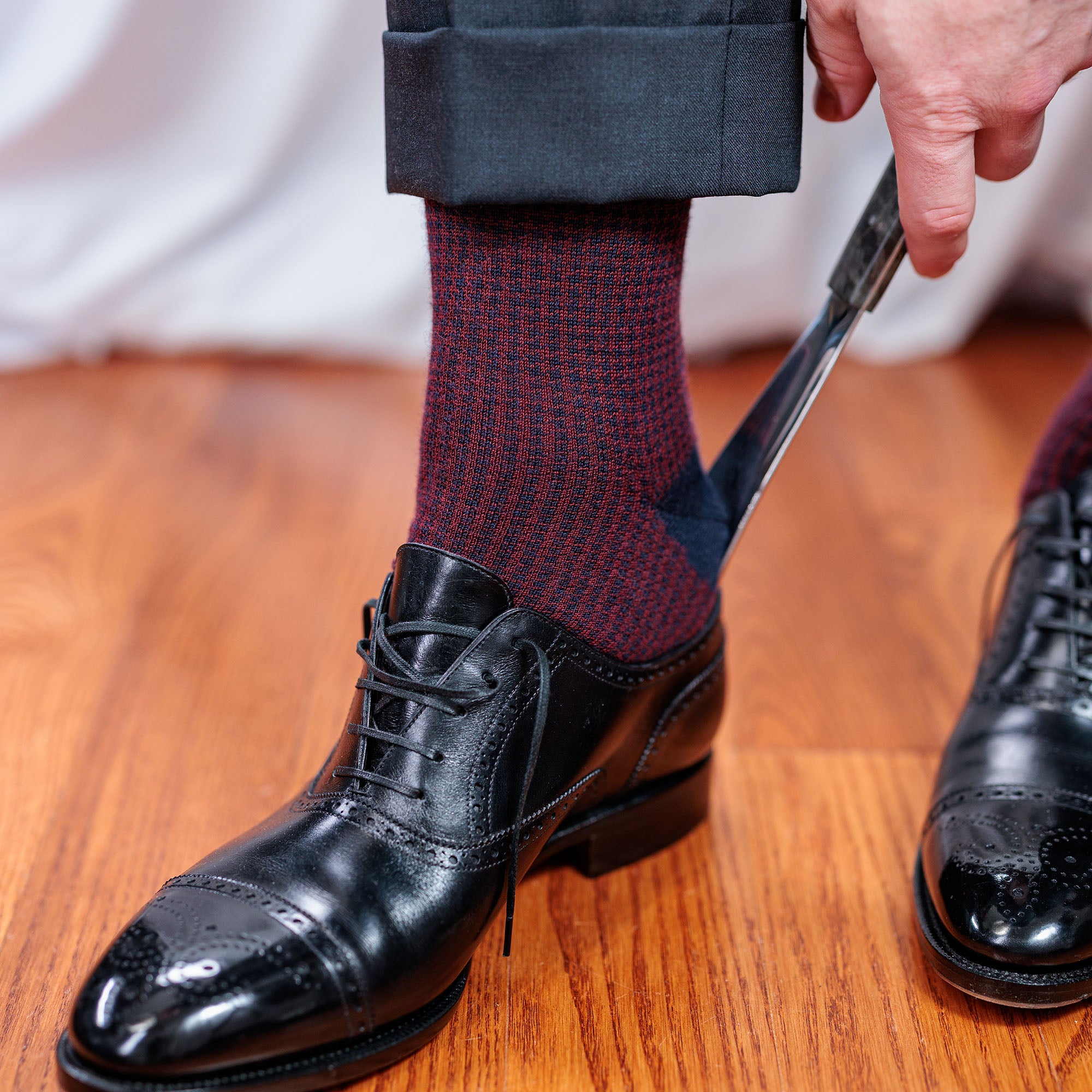 man wearing navy and burgundy houndstooth dress socks with a charcoal suit using a shoe horn to slide into black oxfords