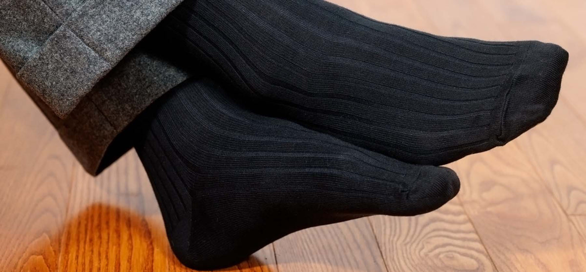 man crossing ankles wearing black ribbed dress socks with grey slacks with a wooden floor in the background
