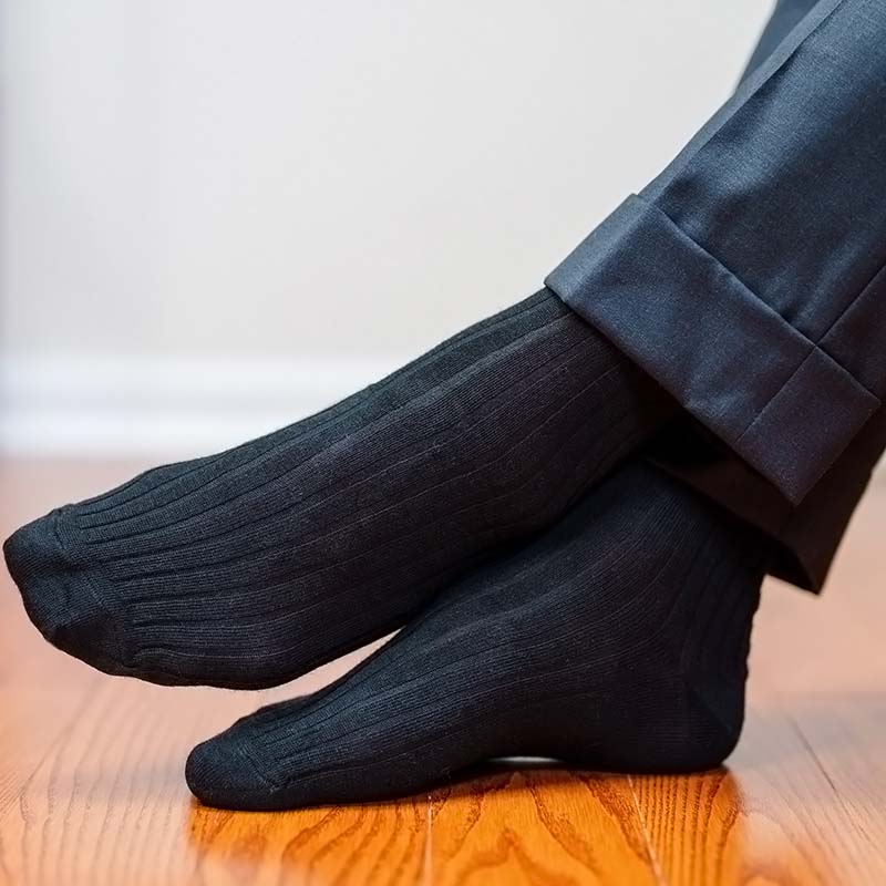 Black ribbed dress socks worn with dark dress pants, shown on a person's feet resting on a polished wooden floor against a neutral indoor background.
