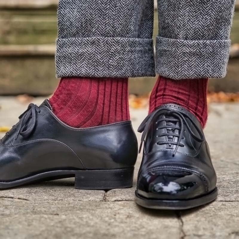 A close-up of a person's feet wearing black leather oxford shoes, dark gray herringbone trousers, and deep burgundy ribbed socks, standing on stone pavement.