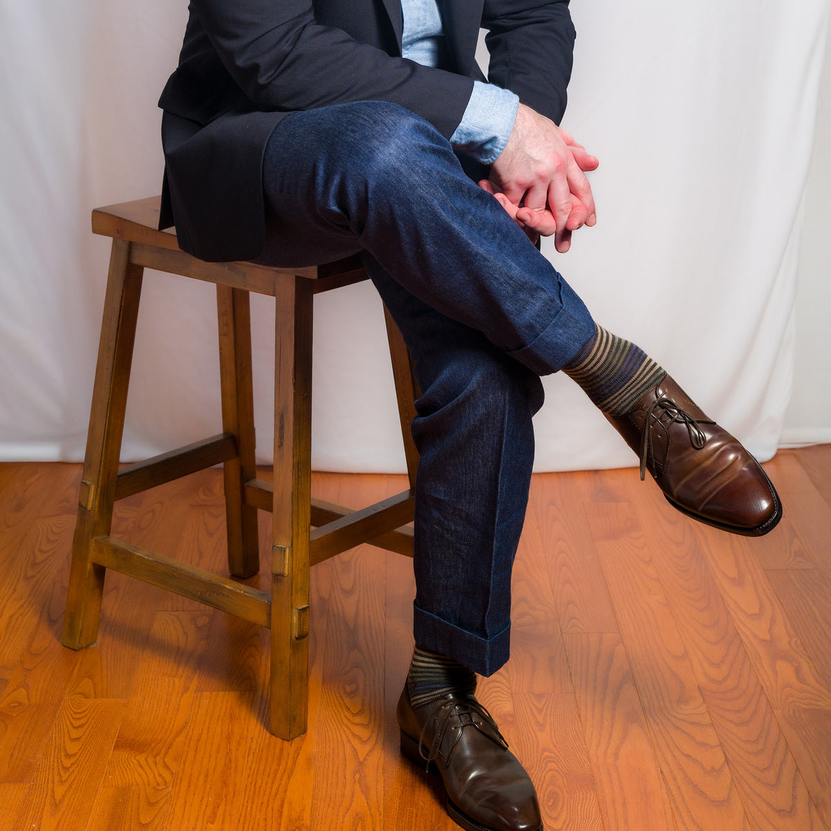 person wearing a suit and brown striped dress socks sitting on a wooden stool with a white curtain background