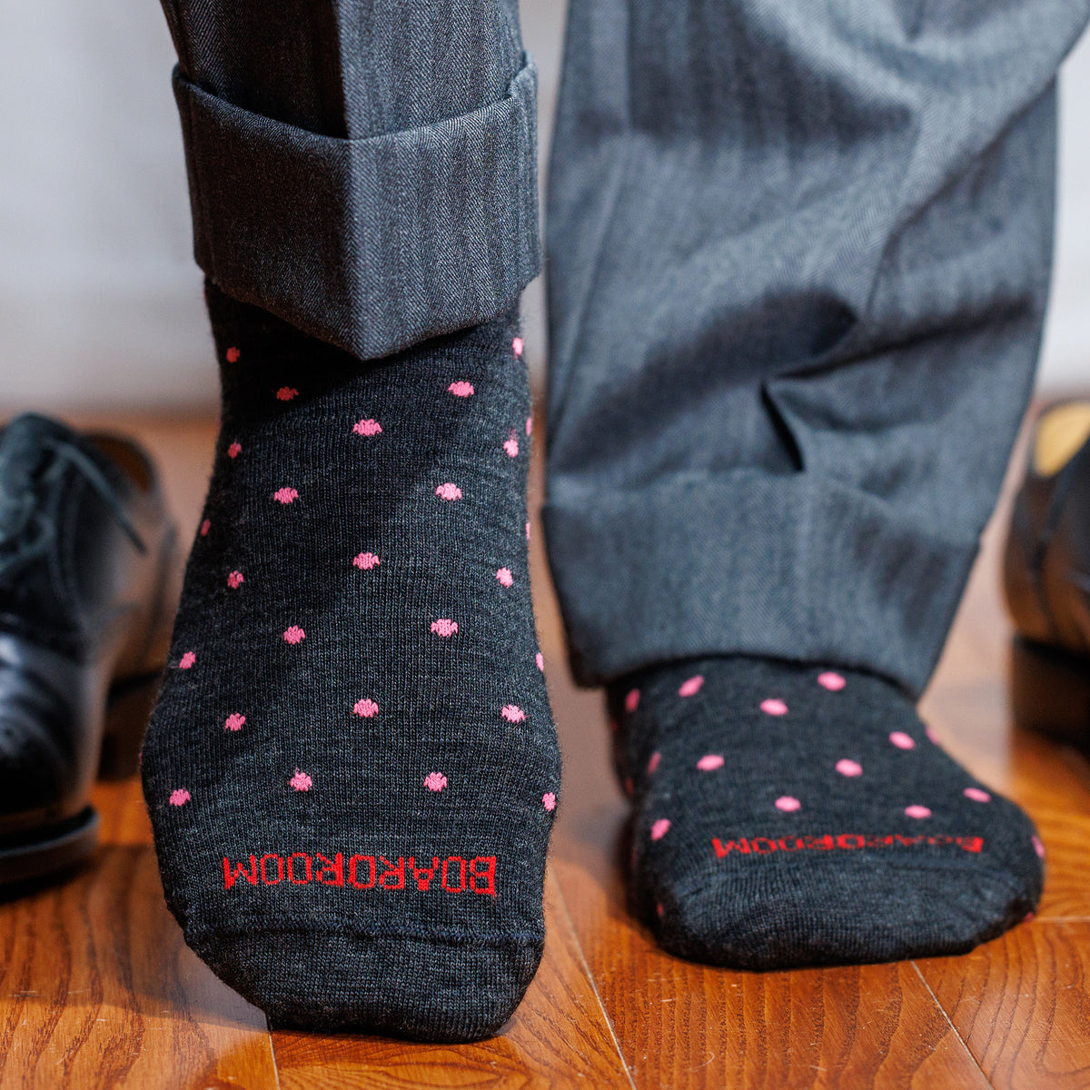 Person wearing dark gray dress socks with pink polka dots and &#39;BOARDROOM&#39; branding on a wooden floor.