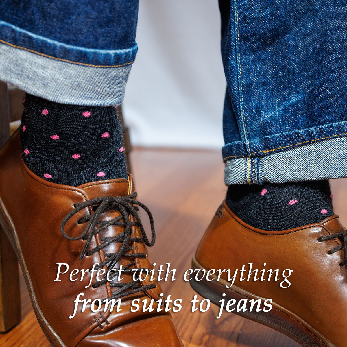 Person wearing charcoal dress socks decorated with pink polka dots, brown leather sneakers, and rolled-up jeans.