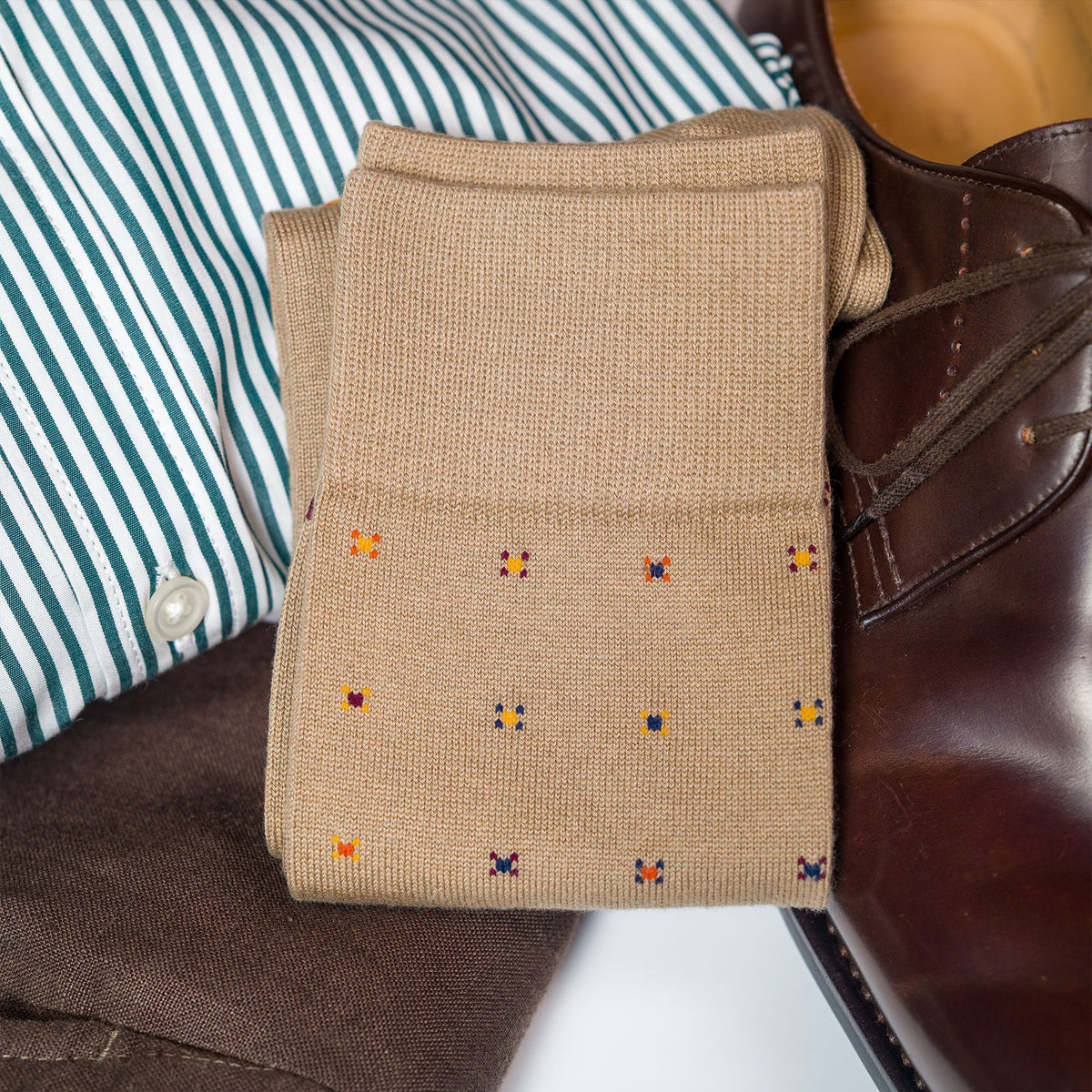a folded pair of khaki geometric pattern dress socks laying on top of clothing and a pair of shoes