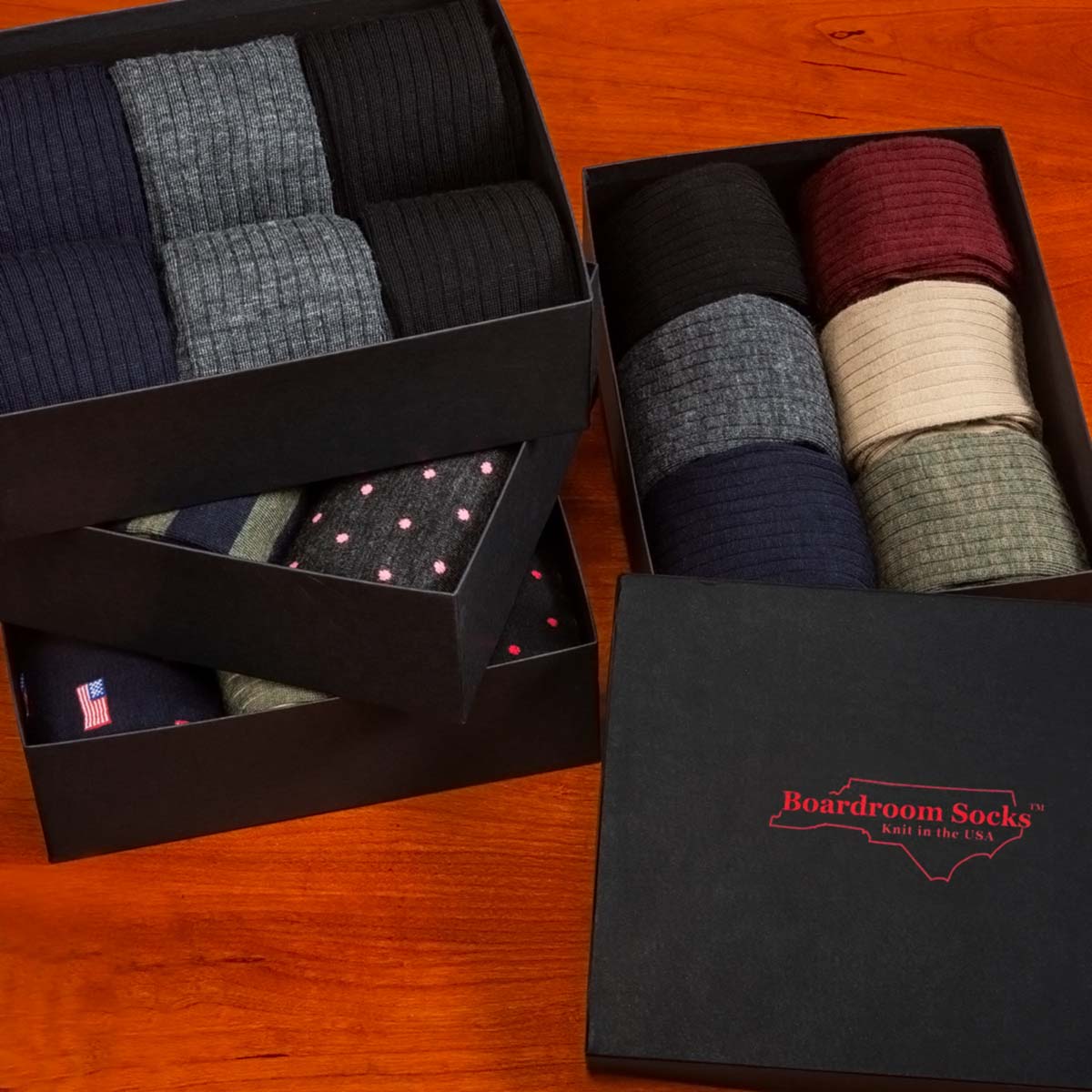 Several black gift boxes of Boardroom Socks on a wooden table, open to show a wide variety of dress socks, and one closed showing the red logo.