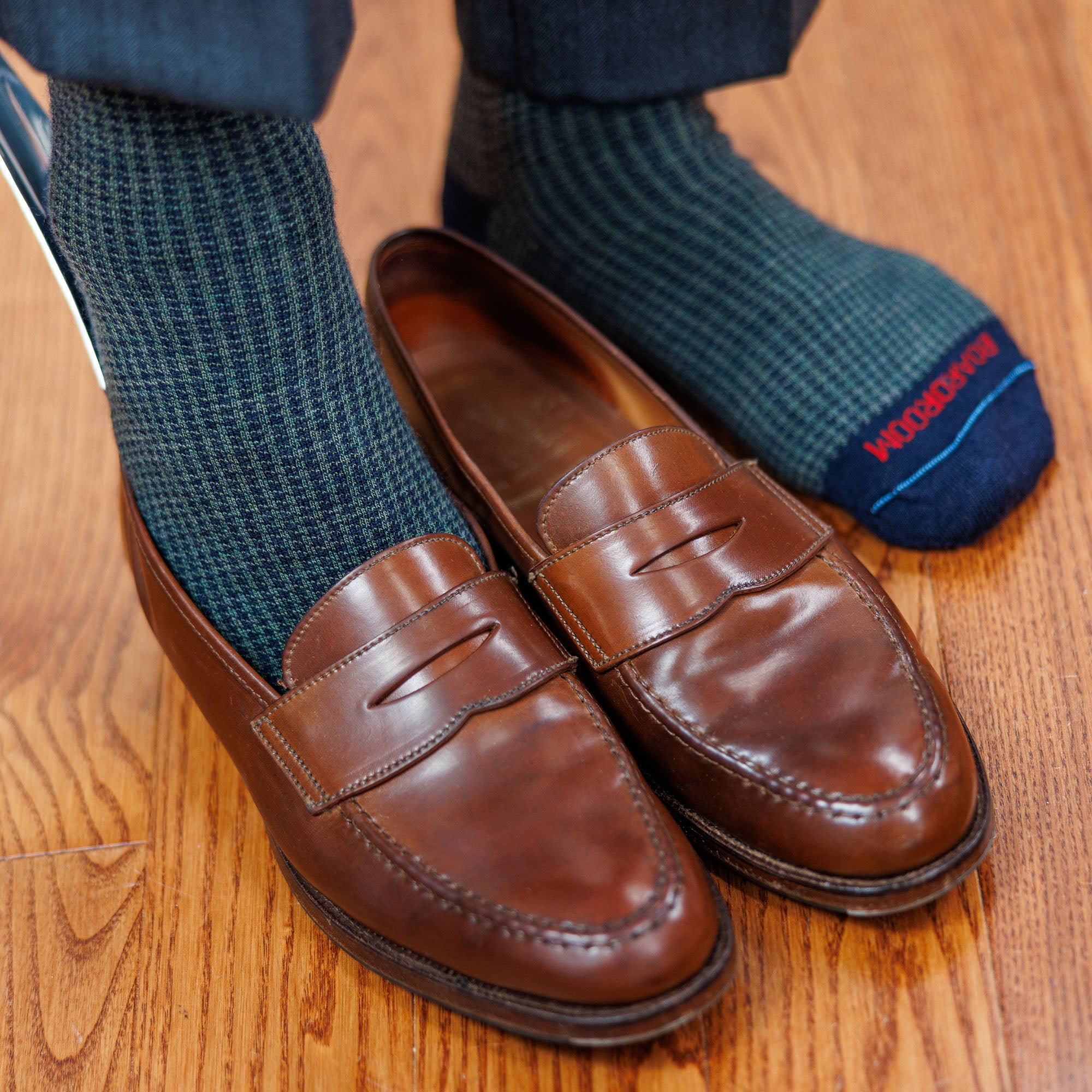 man wearing olive and navy houndstooth dress socks using shoe horn to slide on brown penny loafers