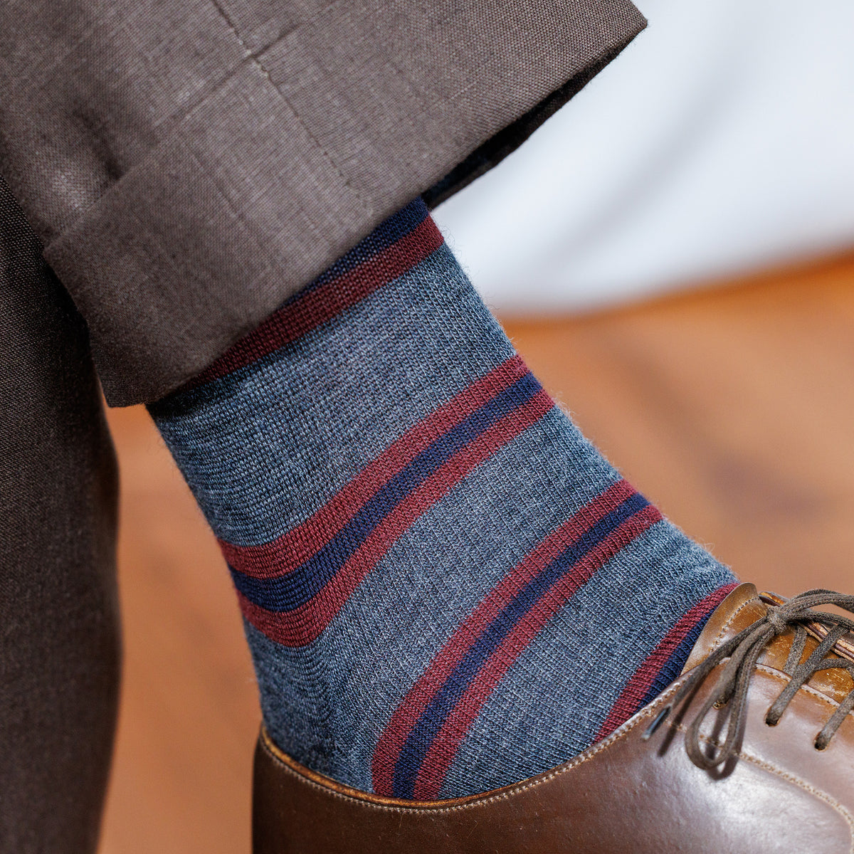 man crossing legs wearing grey wool dress socks decorated with burgundy and navy horizontal stripes paired with brown slacks and brown derby dress shoes