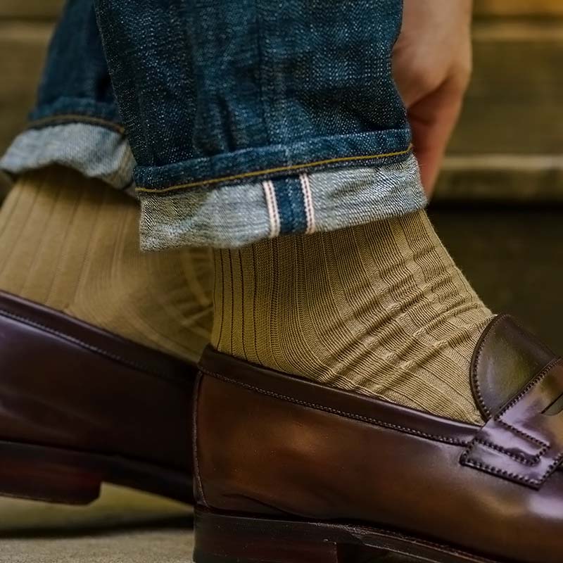 Close-up of a person wearing dark brown leather penny loafers, ribbed khaki cotton dress socks, and cuffed blue jeans.