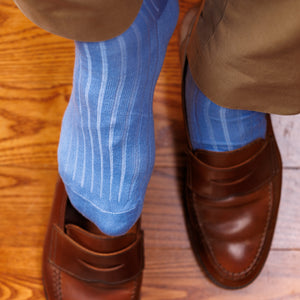 man putting on brown loafers while wearing sky blue ribbed dress socks and khakis