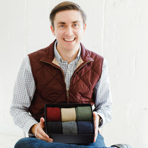man sitting down holding an open gift box containing a variety of colored merino wool dress socks