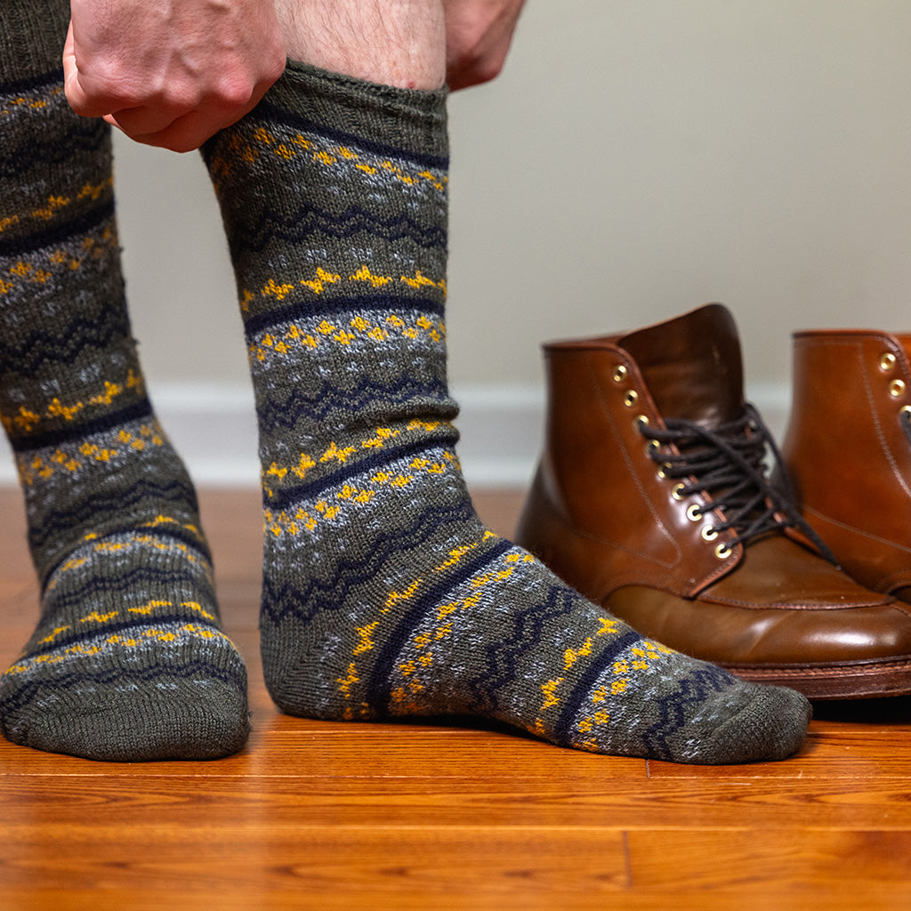 man putting on olive green Fair Isle wool socks while standing on hardwood floor