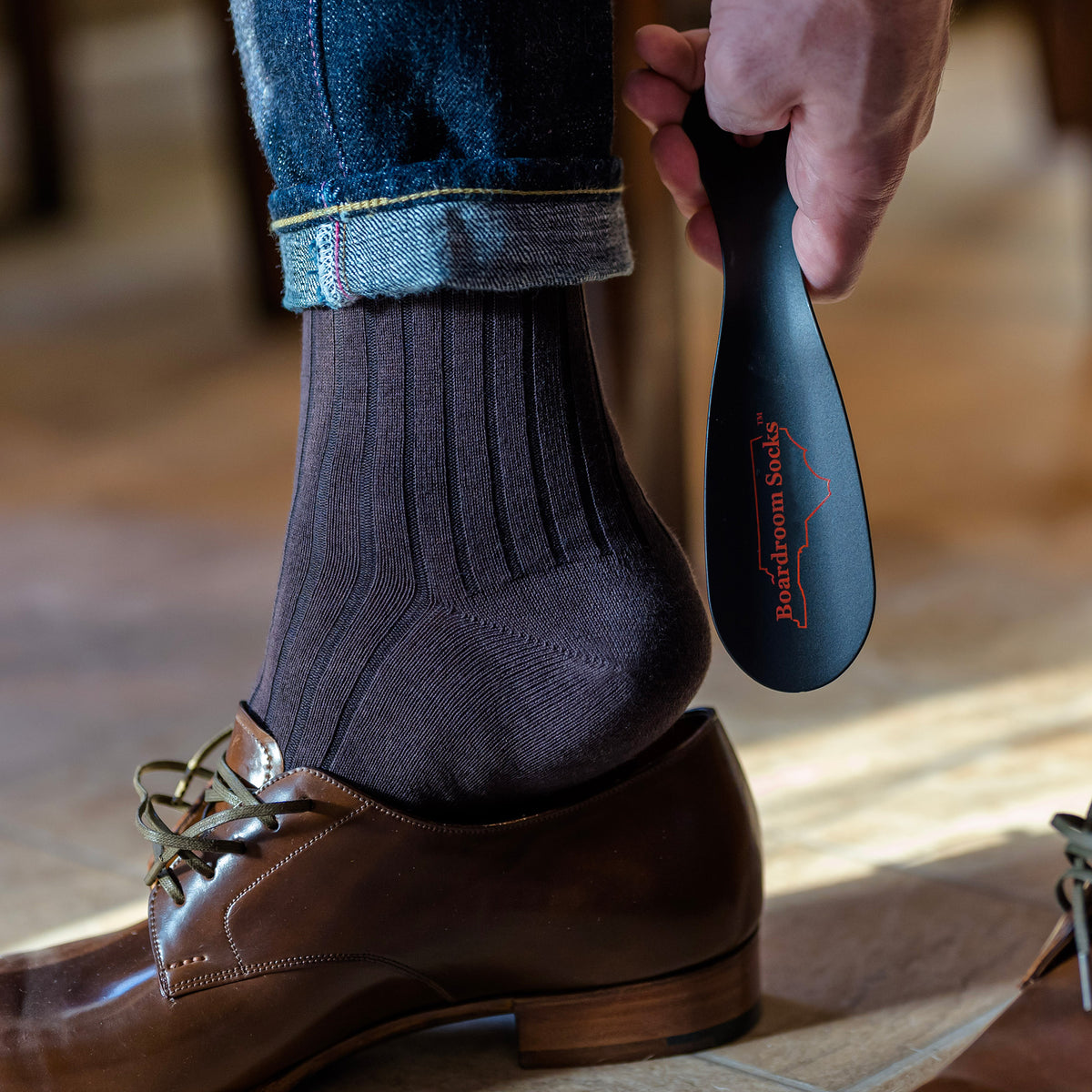 man using black metal shoe horn from Boardroom Socks to slide on brown dress shoes while wearing brown cotton dress socks and jeans