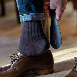 man using black metal shoe horn from Boardroom Socks to slide on brown dress shoes while wearing brown cotton dress socks and jeans
