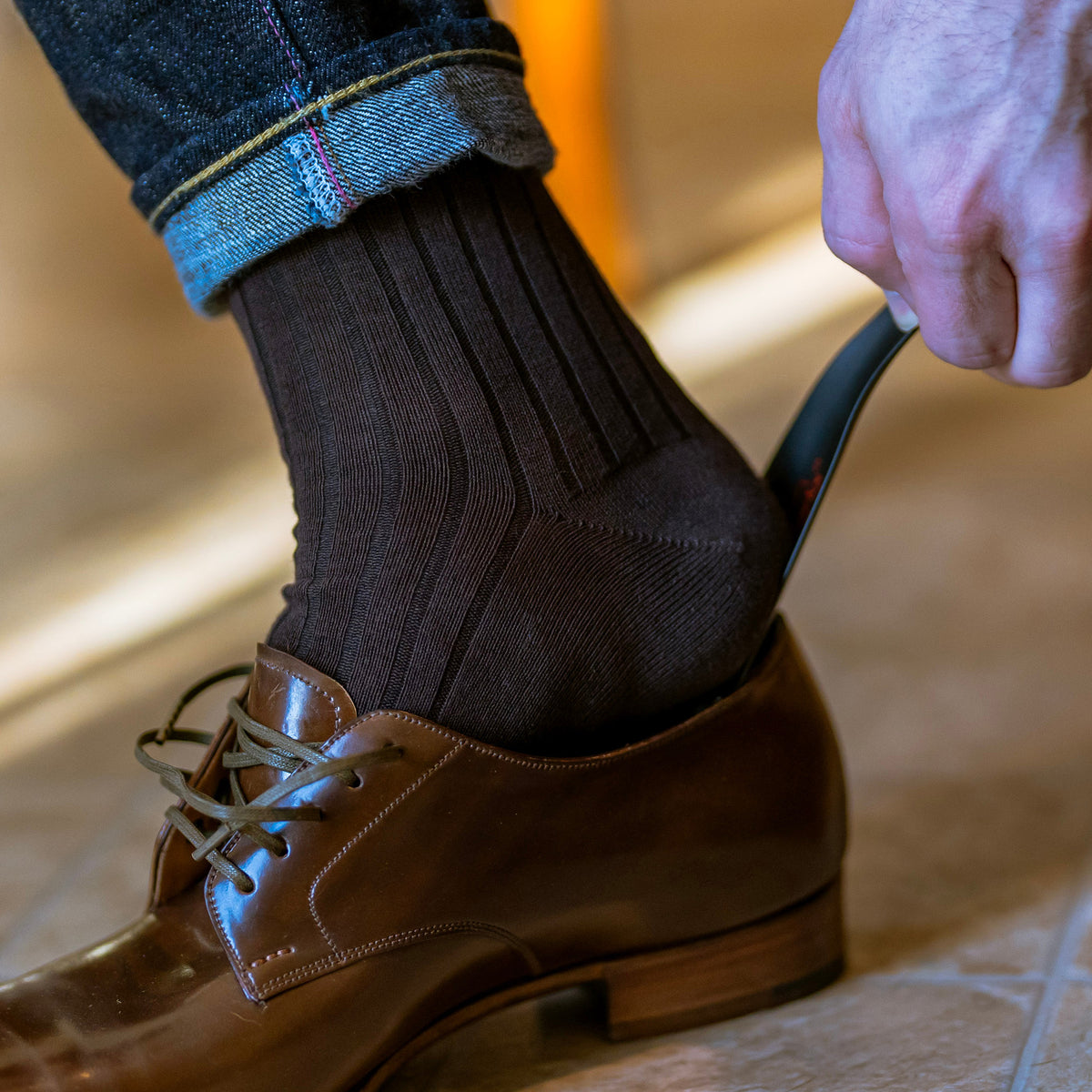 man using metal shoe horn to slide on brown dress shoes while wearing jeans and brown dress socks from Boardroom Socks