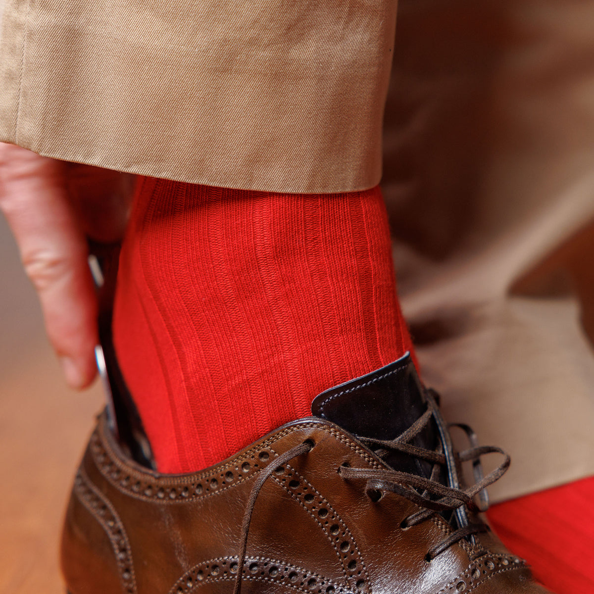 man wearing red dress socks using shoe horn to put on brown wingtip dress shoes