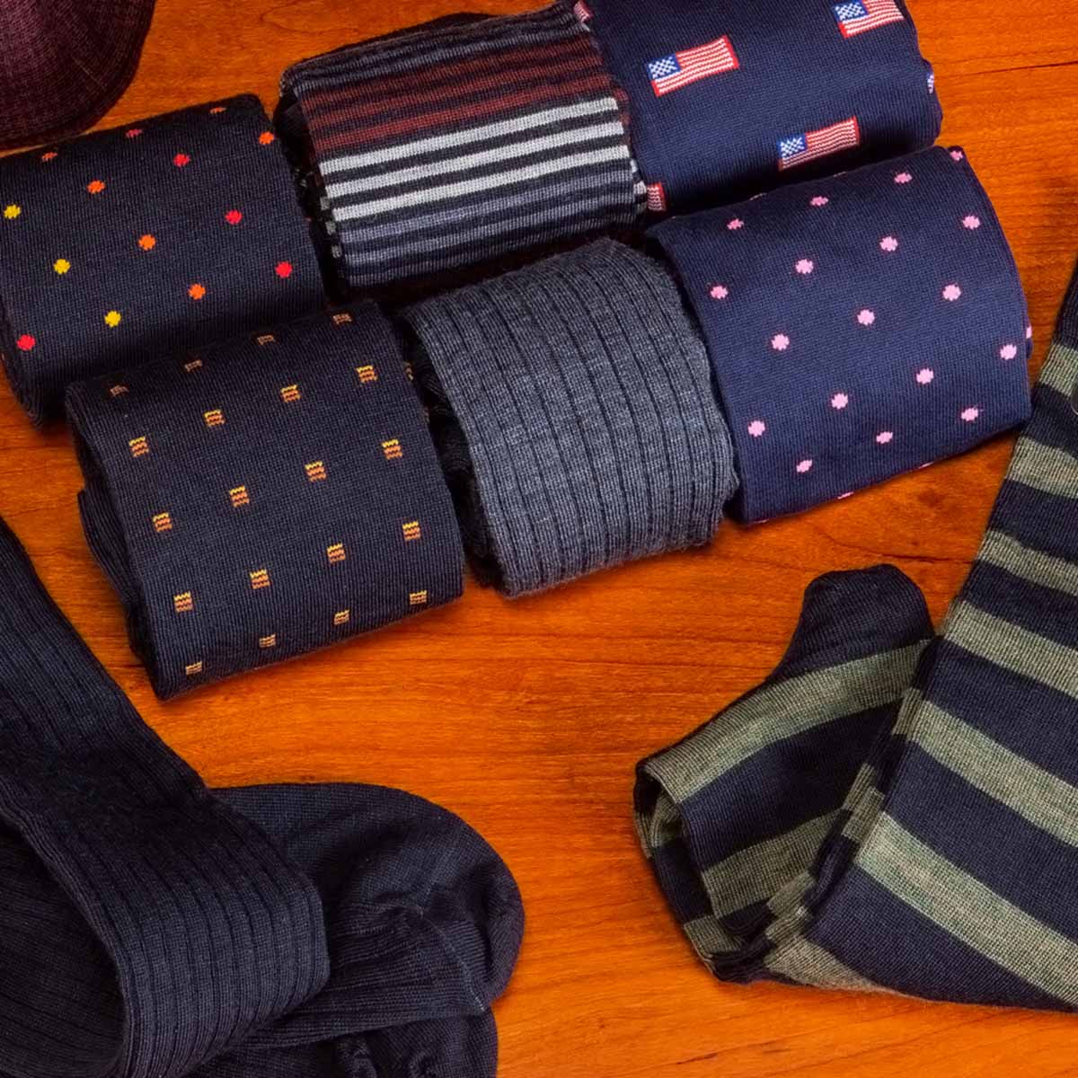 A collection of men's knit dress socks neatly arranged on a warm orange-brown wooden surface. The products feature various patterns including navy blue with pink, yellow, and orange polka dots, navy blue with red and white American flag motifs, navy and gray stripes, and navy and olive green stripes.