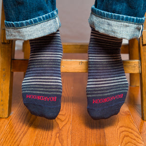 striped dress socks with 'BOARDROOM' branding worn with jeans by a person sitting on a wooden stool