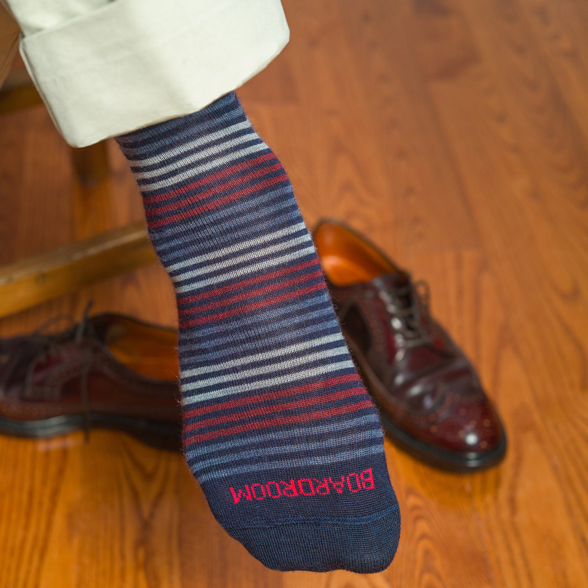 colorful striped dress socks with Boardroom stitched across the toe worn by man sitting on stool with khaki pants