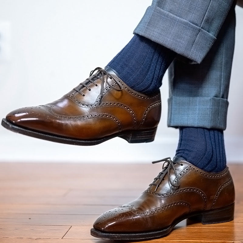 A pair of men's brown leather wingtip oxford shoes with brogue detailing, styled with dark blue socks and cuffed gray trousers on a wooden floor.