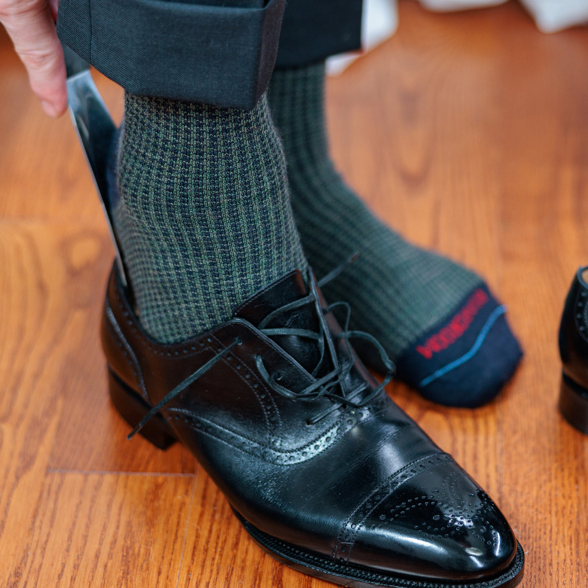 man wearing olive and navy houndstooth dress socks sliding on pair of black oxfords with shoe horn