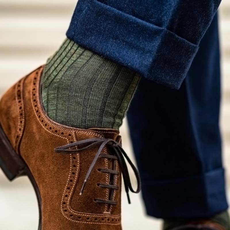 A close-up of a person's foot wearing a brown suede brogue oxford shoe, an olive green ribbed knit sock, and navy blue trousers.