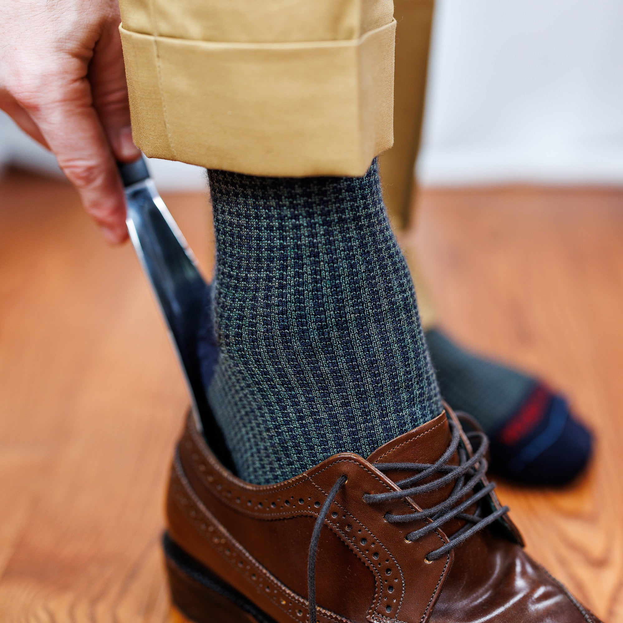 man wearing olive and navy houndstooth dress socks using shoe horn to slide on brown wingtips