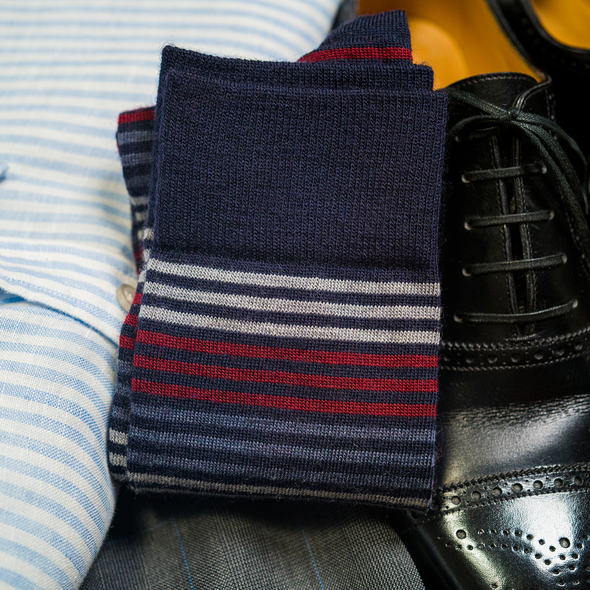 pair of folded striped dress socks sitting atop black dress shoes and a folded striped oxford shirt