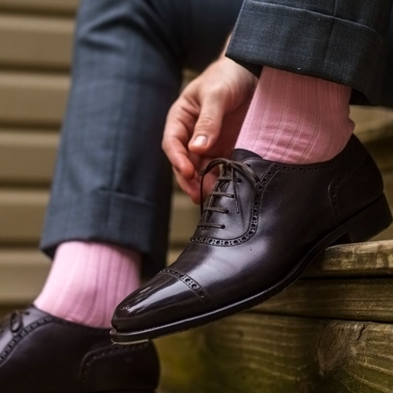 A close-up of a person sitting and tying a dark brown leather oxford dress shoe, wearing light pink ribbed dress socks and dark gray trousers.