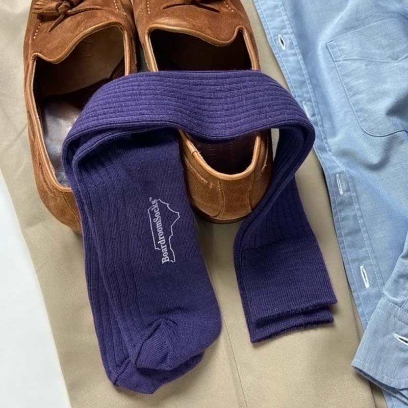 A pair of dark purple ribbed socks with a white 'Boardroom Socks' logo, styled in a flat-lay arrangement with brown suede loafers, a light blue button-up shirt, and beige trousers.
