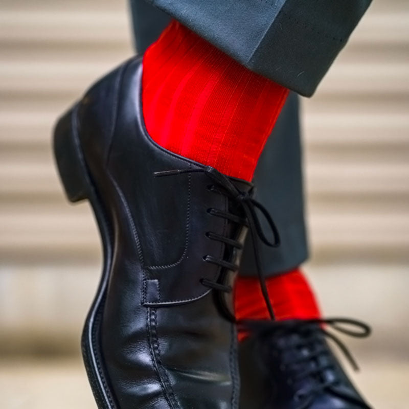 A close-up of a person wearing a black leather lace-up dress shoe with bright red dress socks and dark trousers, shown against a softly blurred beige background.