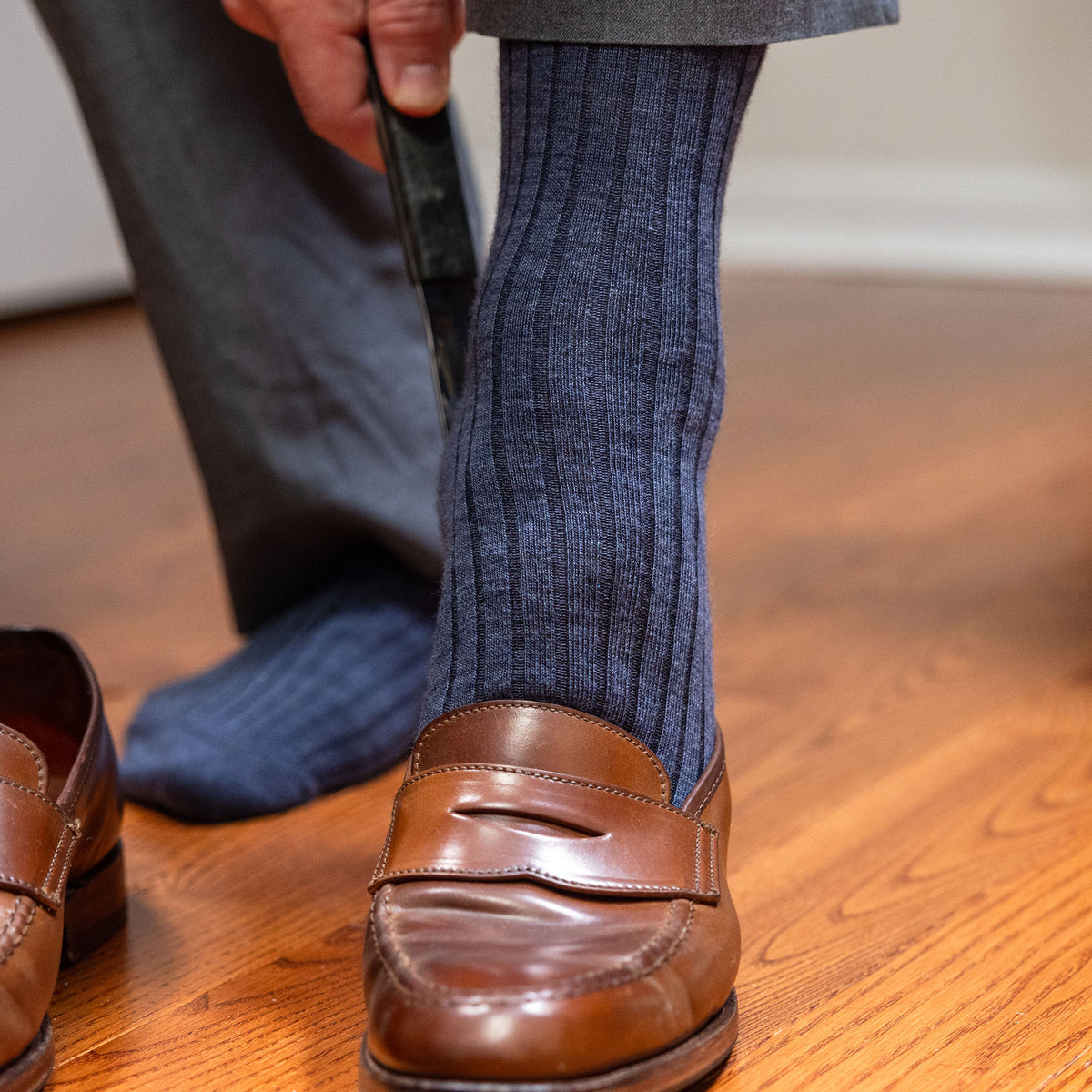 man wearing heather blue wool dress socks using shoe horn to slide on brown penny loafers