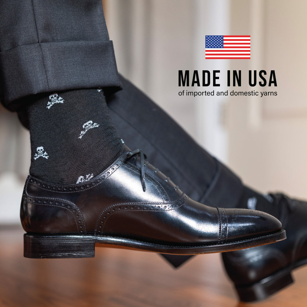 man sitting down crossing legs wearing over the calf merino wool black dress socks with skull and bones pattern with "made in USA" text overlay