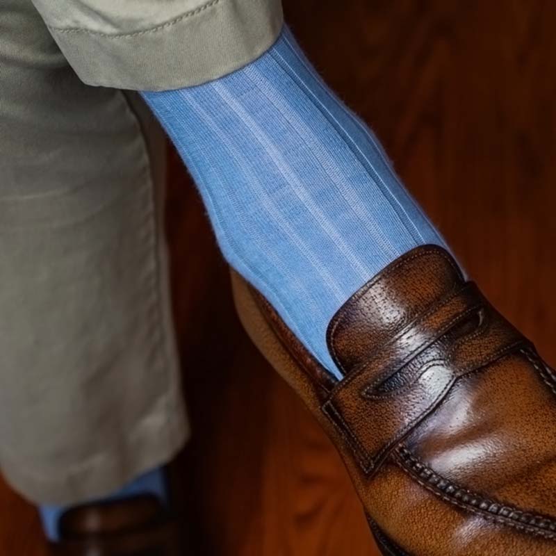 man wearing light blue ribbed dress socks with khakis and brown penny loafers, crossing his legs with a warm wooden floor in the background