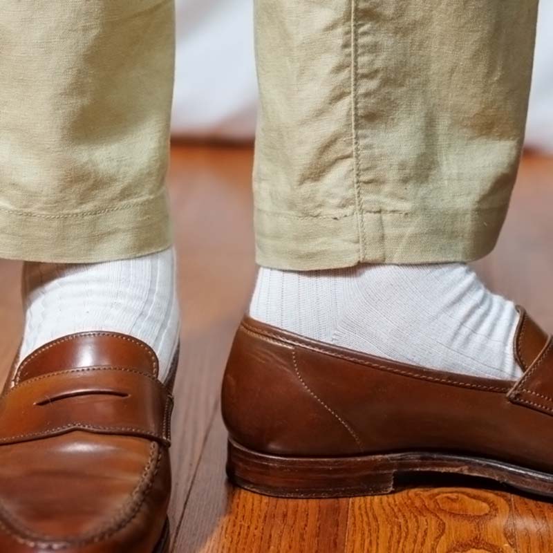 A close-up of a person's feet wearing brown leather penny loafers, white ribbed dress socks, and beige trousers standing on a wooden floor.