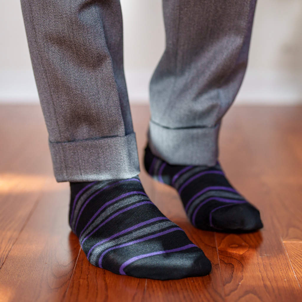 man wearing black purple and grey dress socks with light grey trousers standing on hardwood floor
