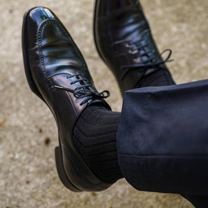 man crossing legs wearing black dress socks with freshly shined black derbies and dark navy trousers