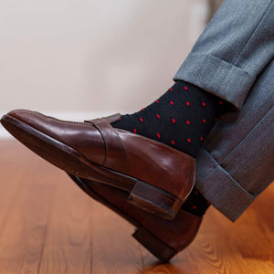 man crossing ankles wearing black dress socks decorated with small red dots paired with burgundy penny loafers and grey trousers